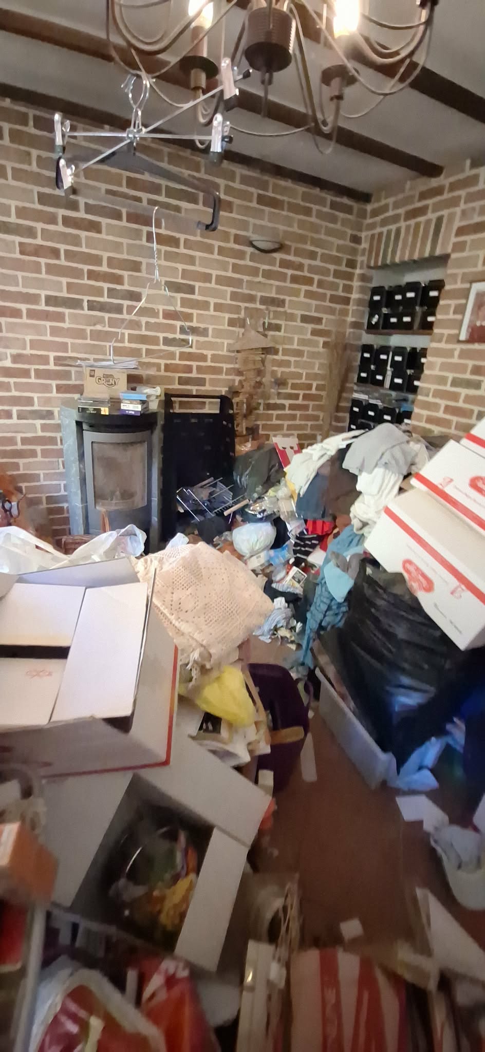 Salon encombré du sol au plafond avant débarras : cartons, vêtements et objets entassés devant un poêle à bois, murs en briques.