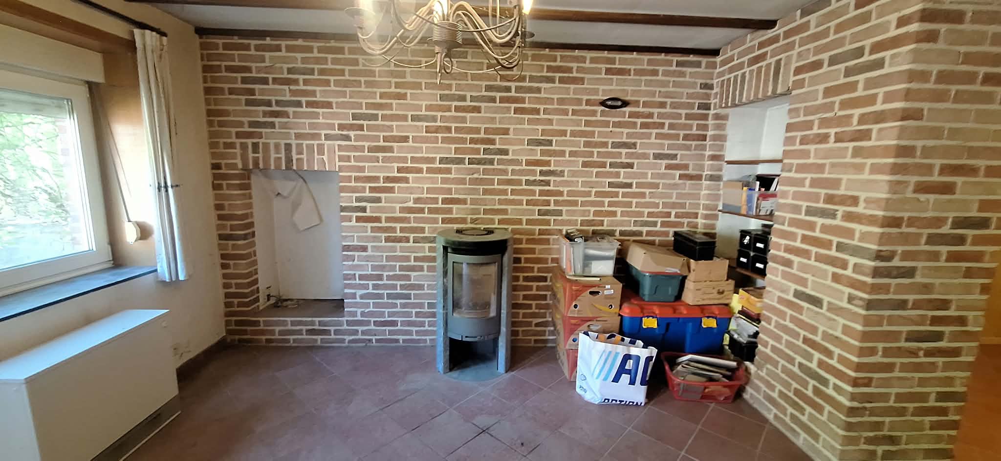 Dezelfde woonkamer na de ontruiming: vrijgemaakte tegelvloer, houtkachel en bakstenen muren zichtbaar, enkele gesorteerde kisten in de hoek.